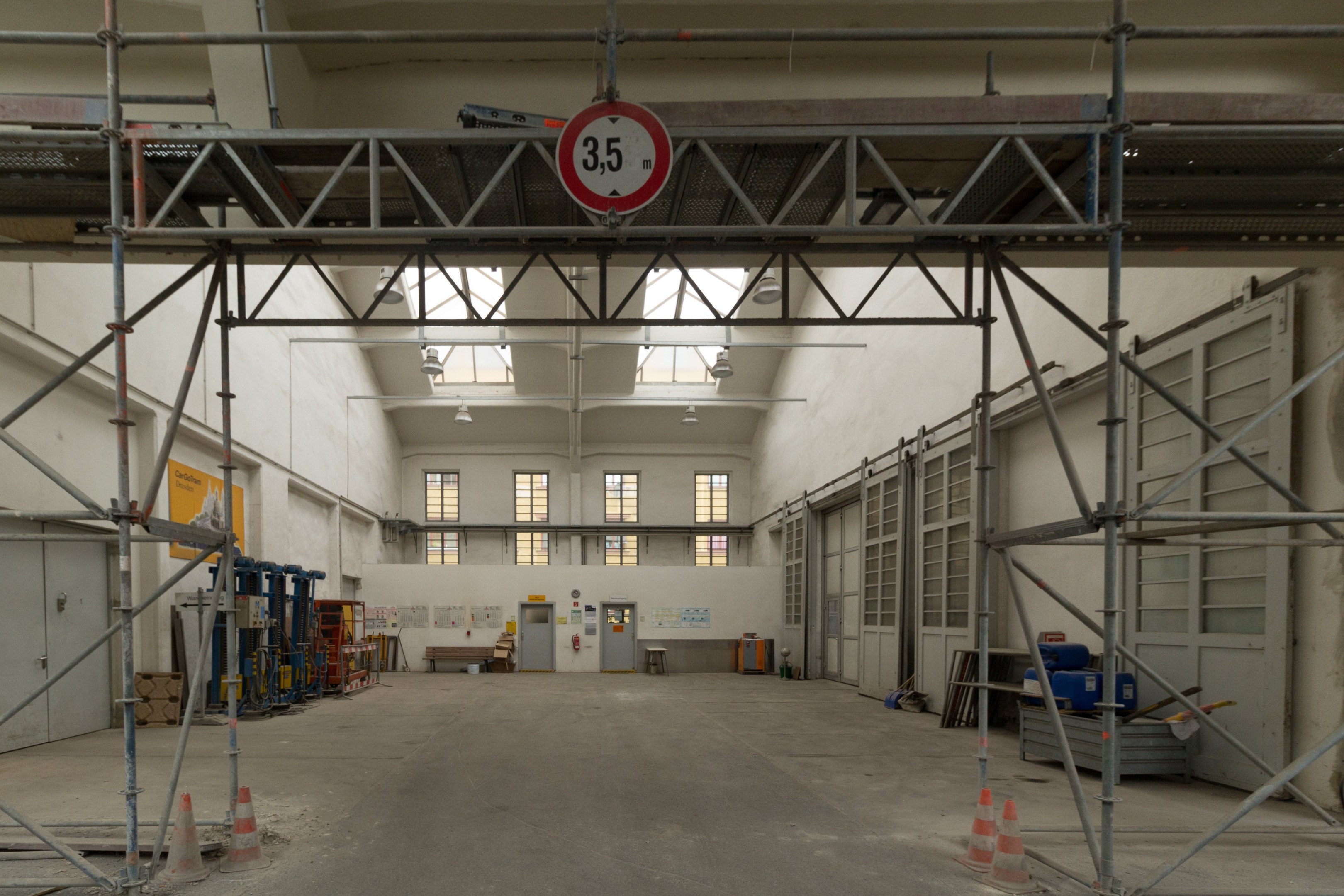 Lagerhalle mit Gerüsten, Verkehrskegeln, Metallstangen, einem Schild, Maschinen, einer Tür, Fenstern, einer Wand, Deckenleuchten und einem Dach.