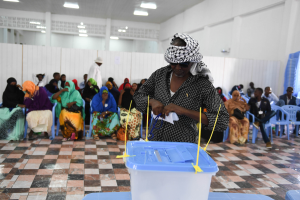 Eine Frau gibt ihre Stimme in einem Wahllokal in Mogadischu, Somalia ab, stehend an einem Tisch mit einer Box, umgeben von sitzenden Menschen und einer stehenden Person im Hintergrund, mit Deckenleuchten, die den Raum erhellen.