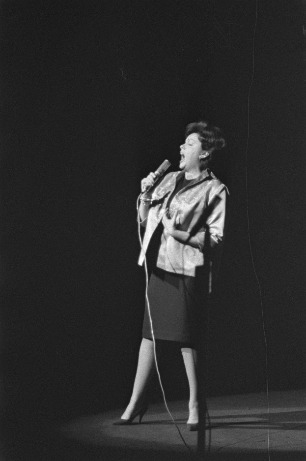 Eine Frau in einem schwarzen und weißen Outfit aus den 1950er-Jahren singt fröhlich auf einer von Scheinwerfern beleuchteten Bühne in ein Mikrofon.