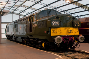Ein Zug befindet sich auf den Schienen neben einer Plattform mit einem Mast auf dem Dach, der Metallstangen mit Lampen trägt.