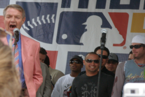 Eine Gruppe von Männern, die meisten tragen eine Schutzbrille, steht und lächelt, wobei einer in einem Blazer in ein Mikrofon spricht; ein weißes, graues und rotes Banner ist im Hintergrund zu sehen.