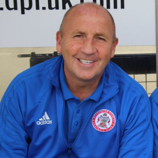 Zwei Männer in einem Raum, einer in einem blauen Jackett neben einem anderen Mann, mit einer Tafel an der Wand dahinter.