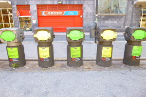 Eine Reihe von Parkautomaten entlang einer Straße mit einem Gebäude, das Glasfenster und eine Rolllade mit Text im Hintergrund sowie einen nahen Müllcontainer zeigt.