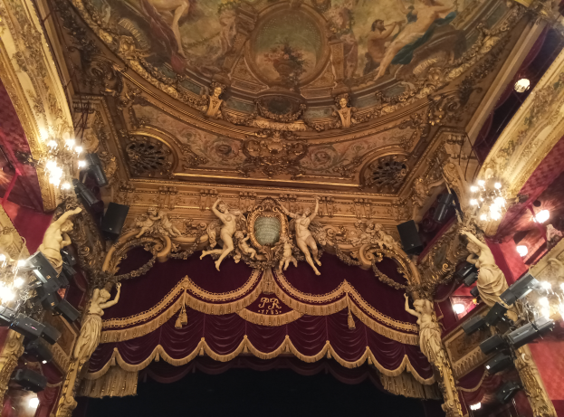 Das Innere eines Theaters mit einem roten Vorhang, Kronleuchtern, Skulpturen, Deckenmalereien und einer Bühne mit ein paar Menschen davor.