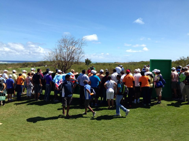 Gruppe von Menschen auf einem grünen Feld bei einem Golfturnier, mit Bäumen, Pflanzen, einem bewölkten Himmel und einem Gewässer im Hintergrund.