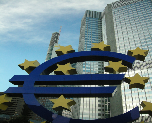Eurozeichen vor dem Europäischen Zentralbank-Gebäude in Frankfurt, Deutschland, mit Bäumen und einem bewölkten Himmel.