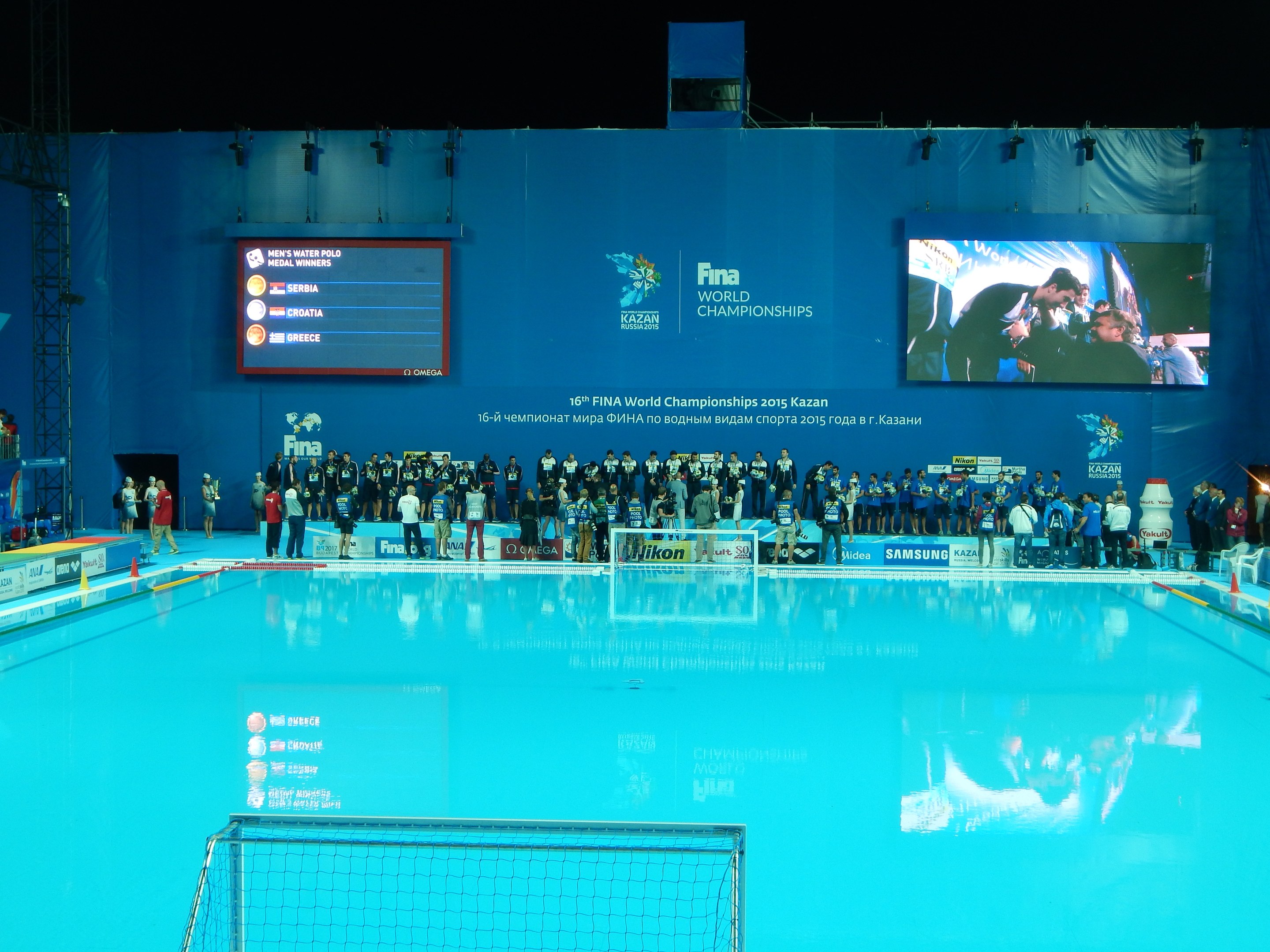 Gruppe von Menschen um ein Hallenbad mit Netz am Boden herumstehend, mit einer Wand im Hintergrund, die den Text und Logos 'FINA World Championships 2015 Kazan' anzeigt.