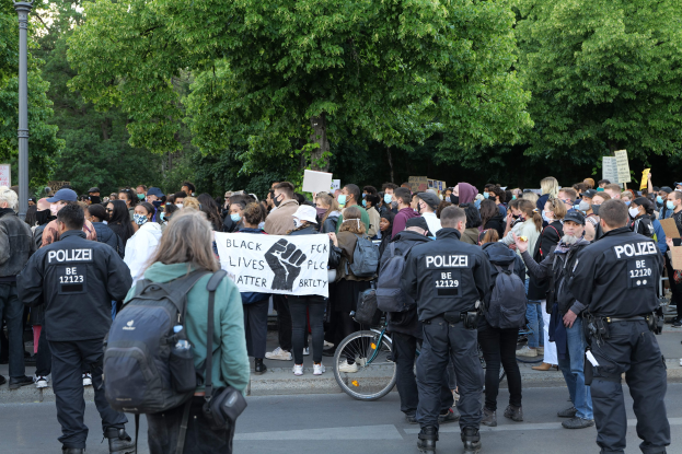 Eine große Gruppe von Menschen steht auf der Straße, einige halten Schilder, andere tragen Kappen und Taschen, mit einem Fahrrad im Vordergrund und Bäumen und einem Mast im Hintergrund, bei einer Black Lives Matter Demonstration in Berlin.