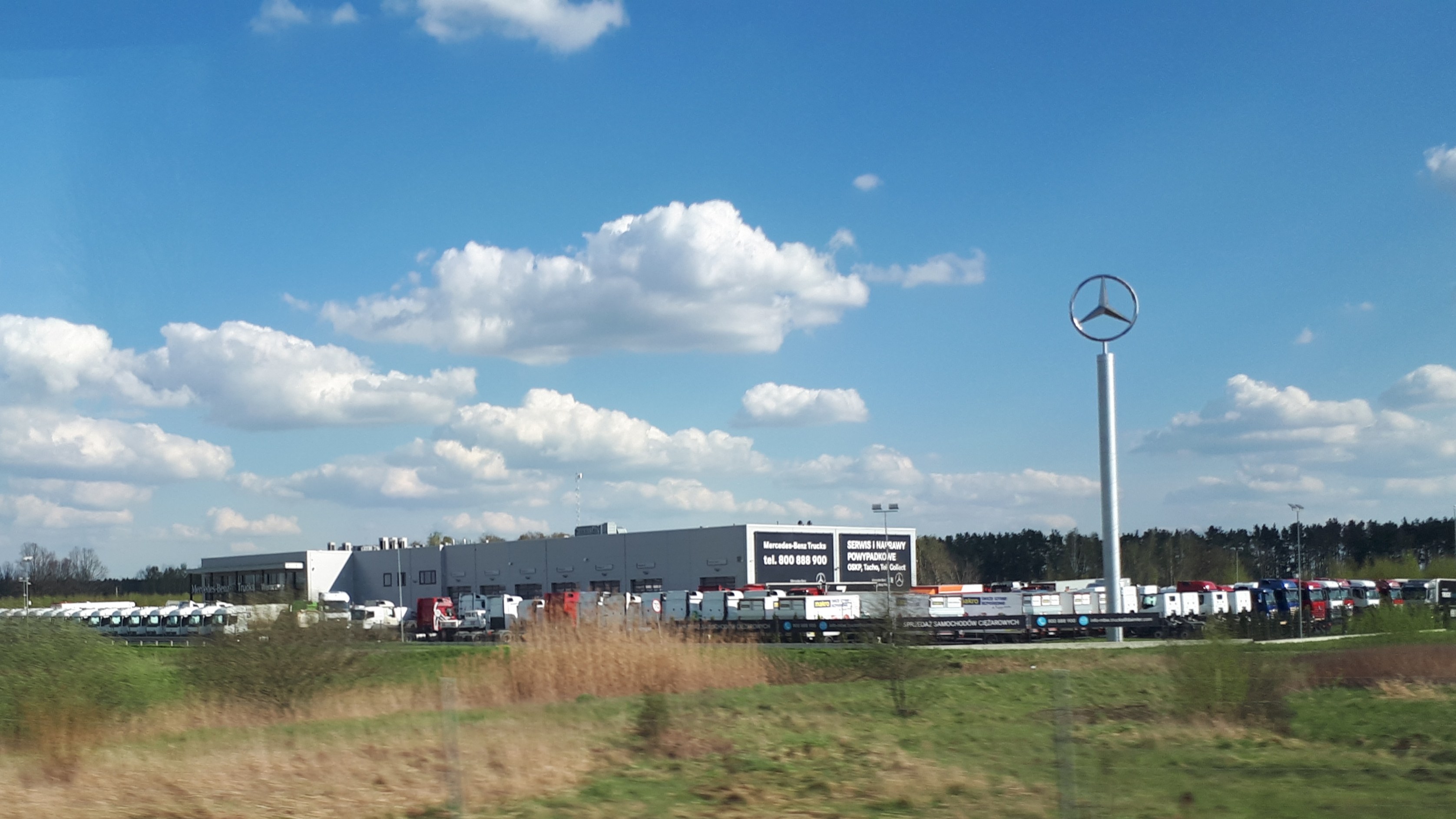 Mercedes-Benz-Fabrik in Deutschland mit Fahrzeugen auf der Straße, Grünflächen, Gebäuden und einem Logo-Mast unter einem bewölkten Himmel.