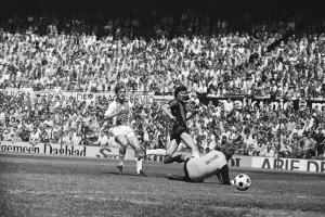 Schwarzes und weißes Foto von Männern, die auf einem Feld Fußball spielen, mit Zuschauern im Hintergrund und einem Bundesliga 1978-79 Ajax 1-1 Hannover 96-Banner.