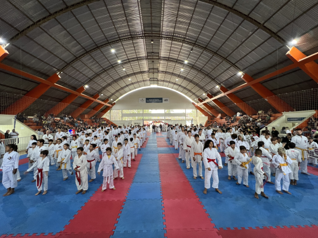 Eine große Gruppe von Menschen auf einer blauen und roten Matte in einer Turnhalle mit anderen auf Treppen sitzend, unter Deckenlampen, mit Hintergrundtafeln, die ein Taekwondo-Event anzeigen.