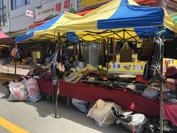 Ein Markt mit Zelten, die verschiedene Gegenstände wie Taschen und Hüte präsentieren, vor Häusern mit Fenstern und Texttafeln im Hintergrund.
