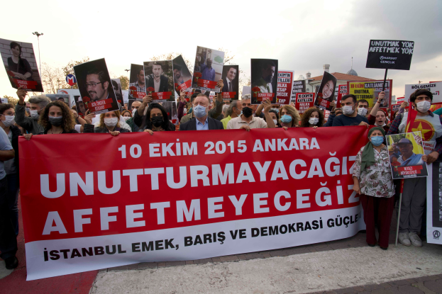 Eine Gruppe von Menschen mit Masken und Schildern und Transparenten protestiert vor einem Gebäude in Ankara, Türkei, mit Bäumen, Laternenmasten und einem klaren Himmel im Hintergrund.