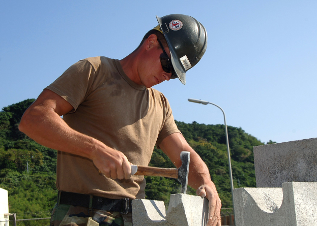 Ein Mann in einem Helm und einer Schutzbrille verwendet einen Hammer, um eine Wand im Freien mit Bäumen, einem Laternenmast und einem klaren blauen Himmel im Hintergrund zu bauen.