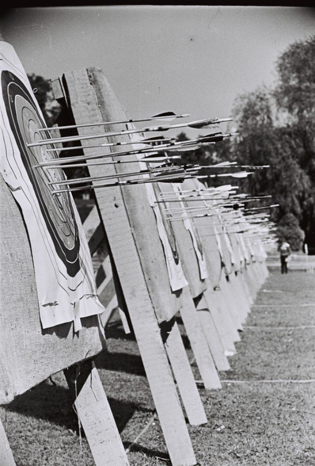 Schwarzes und weißes Foto eines Bogenschießziels mit Pfeilen in der Mitte, umgeben von Bäumen und einer Person im Hintergrund, mit sichtbarem Boden unten.