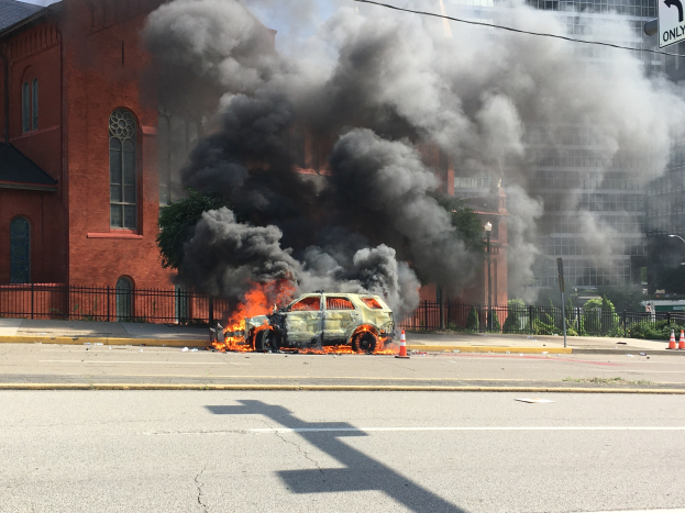 Auto steht in Flammen am Straßenrand mit Rauch, der aus ihm aufsteigt, umgeben von Gebäuden, Bäumen und anderen urbanen Elementen unter einem klaren Himmel.