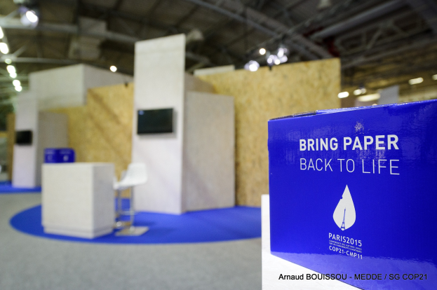 Messestand mit einem blauen und weißen "Bring Paper Back to Life"-Schild auf der rechten Seite, das einen blauen Stuhl, einen Tisch, einen wandmontierten Fernseher und eine Deckenbeleuchtung im Hintergrund zeigt.