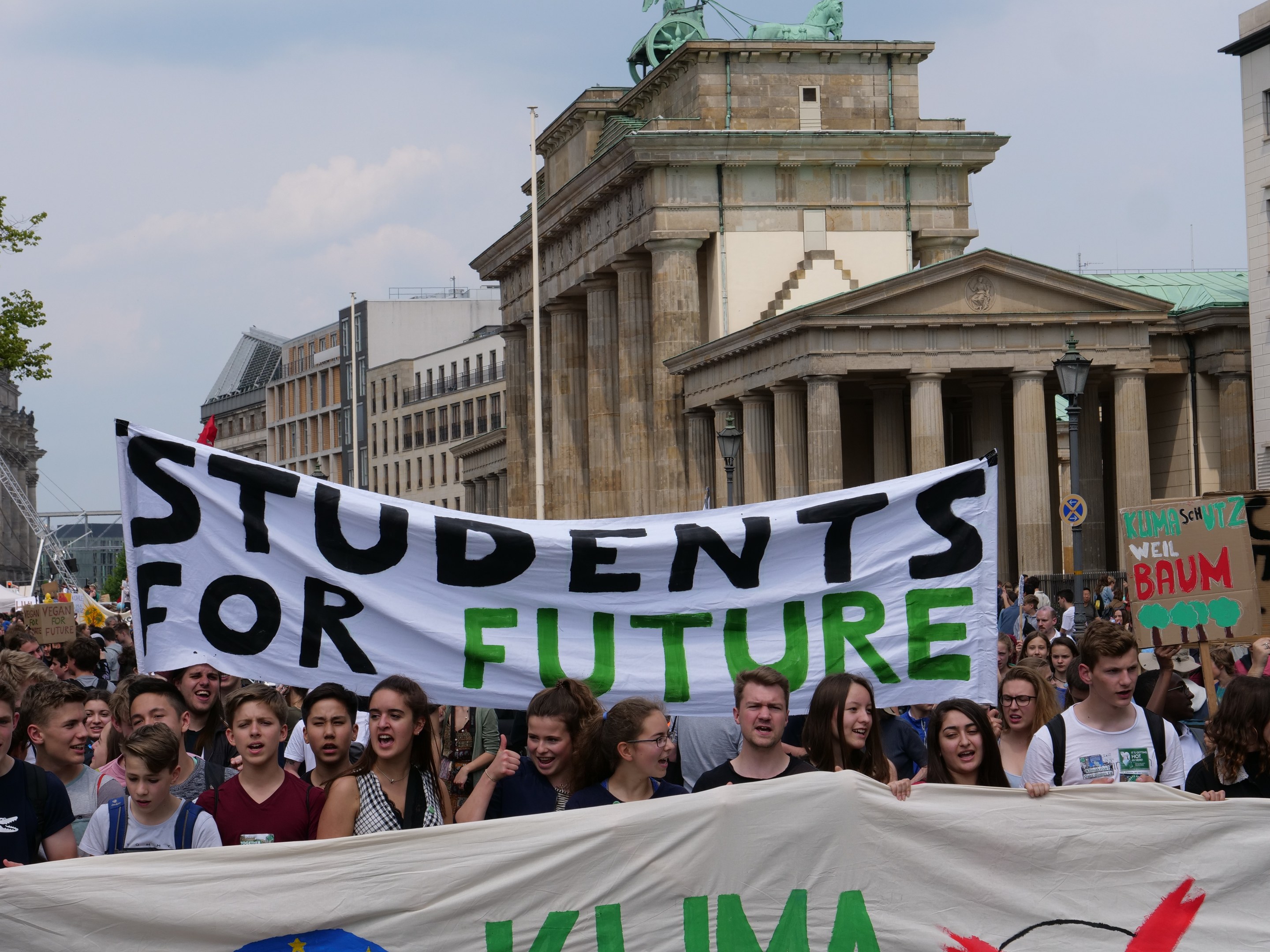 Gruppe von Schülern marschiert in Berlin mit einem bunt bemalten 'Students for Future' Schild gegen eine Kulisse aus Gebäuden, Bäumen und Himmel.