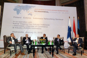 Eine Konferenzbühne mit Menschen, Stühlen, einem Mikrofon, einem Tisch, Flaschen, Blumen, Gläsern und anderen Gegenständen, mit Flaggen und einem Banner in der Mitte und einem Gebäude mit Vorhängen im Hintergrund.