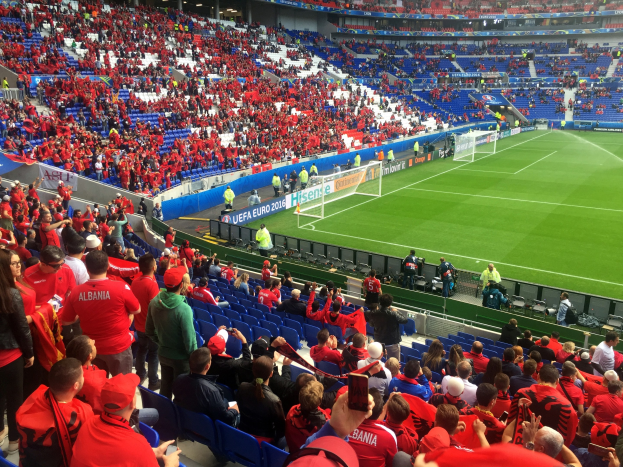Eine große Menschenmenge in einem Stadion schaut einem Fußballspiel zu, mit Menschen, die auf Stühlen sitzen und stehen, Bannern, einem Torpfosten und Deckenbeleuchtung.