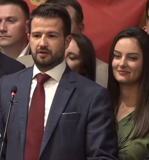 Ein Mann im Anzug spricht vor einem Publikum mit Mikrofon an einem Rednerpult, hinter einem Banner, wahrscheinlich über ein Regierungssozialmedienverbot diskutierend.