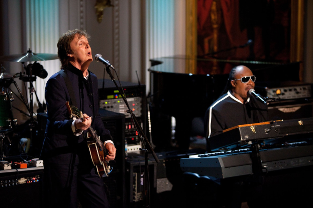 Paul McCartney spielt eine Gitarre und singt in ein Mikrofon, während Stevie Wonder eine Piano spielt und eine Brille trägt auf der Bühne bei den Grammys, mit Musikinstrumenten und gerahmten Kunstwerken im Hintergrund.