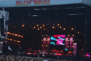 Eine B√ľhne mit Lichtern, einem Bildschirm und Musikern, die Instrumente spielen, mit einem Banner oben, einer Menge unten und Fahnen mit St√§ben und Luftballons.