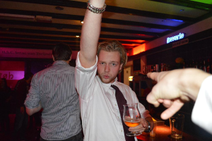 Eine Gruppe von Menschen, die in einer Bar stehen, mit einem Mann in einem weißen Hemd und schwarzer Krawatte, der ein Weinglas in der Nähe der Bartheke hält, auf der viele Weinflaschen und "Bavaria" stehen.