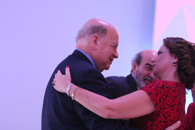 Zwei Menschen umarmen sich herzlich auf einer Bühne, umgeben von anderen, und einer trägt ein Armband.