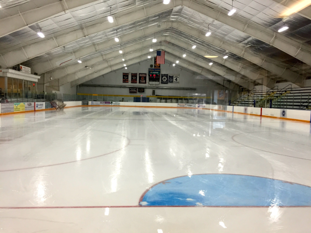 Große Indoor-Eisbahn mit Deckenbeleuchtung, Werbetafeln, Treppen mit Geländern und einigen Menschen in einer Sportanlage.