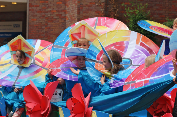 Eine Gruppe von Kindern in bunten Kostümen fährt auf einem Umzugswagen, hält Schirme und hat ein Gebäude, Pflanzen und Lichter im Hintergrund.