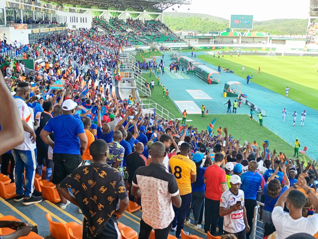 Eine große Menschenmenge versammelt sich vor einem eingezäunten Fußballfeld mit Hügeln und einem klaren blauen Himmel und wartet gespannt auf den Beginn eines Spiels während der 2019 African Cup of Nations.