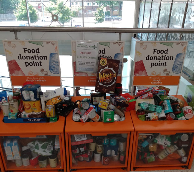 Orange Schübe in einem mit gespendeten Lebensmitteln gefüllten Gebäude, mit Texttafeln und einem Blick durch Glasfenster auf Fahrzeuge, Bäume, Gebäude und Himmel.