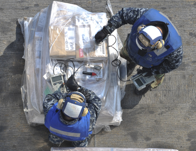 Zwei Männer in blauen Uniformen und Helmen arbeiten an einem großen Drohnenmodell, halten Gegenstände, während eine Plastikabdeckung auf dem Boden liegt.