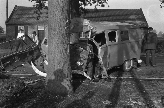 Eine schwarz-weiße Szenerie eines verunglückten Autos am Straßenrand mit einem Baum im Vordergrund, einigen Menschen in der Nähe und Häusern, Bäumen und einem Mast im Hintergrund.