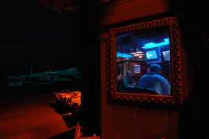 Ein Mann in einem blauen Hemd sitzt in einem schwach beleuchteten Raum und schaut ernst auf einen Fernseher vor ihm, mit ein paar Gegenständen im Hintergrund.