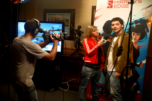 Ein Mann mit einer Videokamera und einem Headset filmt ein Interview zwischen einem Mann und einer Frau, mit einem Fernseher und einem gerahmten Foto im Hintergrund.