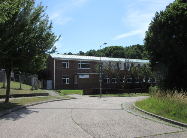 Ein Schulgeb├Ąude mit Fenstern, einer ├ťberspannung, einem Verkehrsschild, einer Gruppe von B├Ąumen, Gras, einem Zaun und einem bew├Âlktem Himmel, das in der Mitte eines Parks umgeben von saftigem Gras liegt.