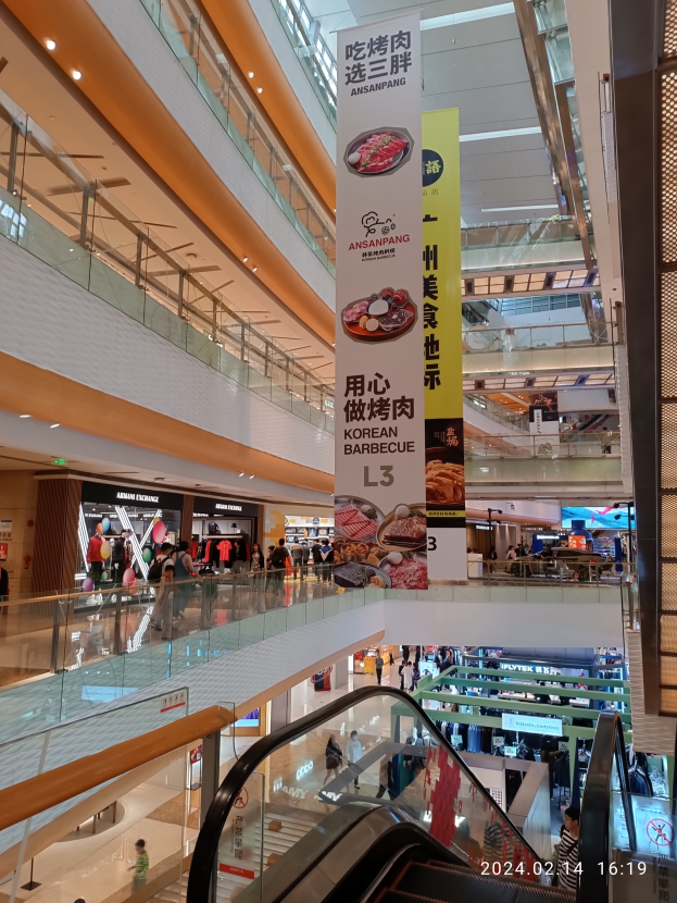 Ein Blick ins Innere eines Einkaufszentrums mit einer Rolltreppe in der Mitte, Menschen, die herumlaufen, einige mit Handtaschen, und Schilder mit Text an der Decke, beleuchtet von Lampen mit Geschäften im Hintergrund.