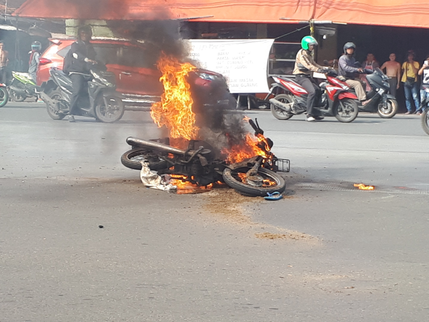 Motorrad in Flammen an der Straße mit einem Auto und einem Van im Hintergrund, umgeben von Menschen in Helmen und anderen, mit einem Banner und einem Zelt im Bild.