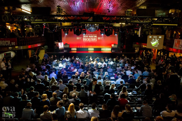 Eine große Menschenmenge sitzt auf Stühlen vor einer Bühne, die von Scheinwerfern beleuchtet und von Geländern umgeben ist, mit Bildschirmen und Bannern mit Text auf der Bühne und einem Wasserzeichen in der linken unteren Ecke, das eine Atmosphäre der Aufregung und Vorfreude für ein Online-Marketing-Rockstars-Event schafft.