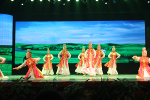 Eine Gruppe von Menschen in farbenfrohen Kostümen tritt auf einer Bühne auf, die von Scheinwerfern überkopf beleuchtet wird und von Pflanzen umgeben ist, mit einem Bildschirm im Hintergrund.