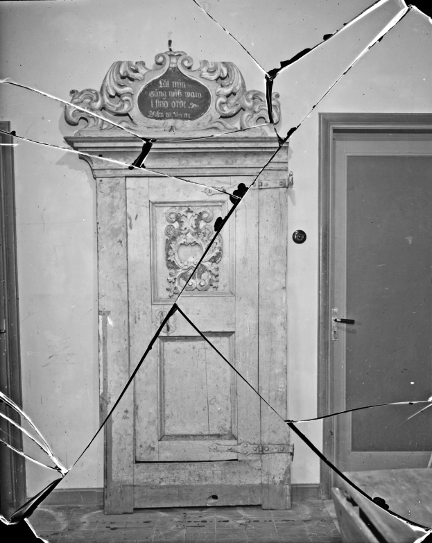 Ein Schwarz-Weiß-Foto einer kaputten Tür mit verstreuten Glasscherben in einem Raum mit einer sichtbaren Wand im Hintergrund.