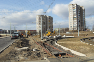 Eine Baustelle mit einer Bahnschiene, Geb├Ąuden, Stra├čenmasten, Stra├čenlaternen, Strommasten, Stromkabel, Kraftfahrzeugen, Personen, Steinen, B├Ąumen und einem bew├Âlktem Himmel.