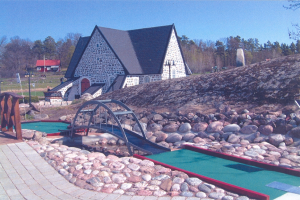 Ein Minigolfplatz mit einem Haus im Hintergrund, umgeben von Felsen, einer Brücke, einem Weg, Gras, Pflanzen, Bäumen und anderen Gegenständen, unter einem sichtbaren Himmel.