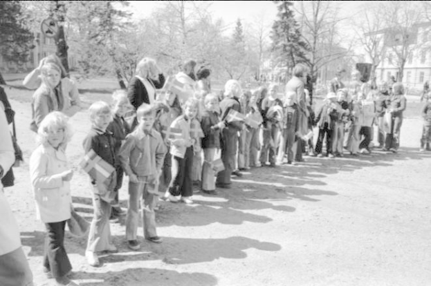 Schwarz-weißes Bild einer Gruppe von Menschen, die während einer Protestdemo auf dem Schulgelände mit Flaggen in einer Reihe auf einem Schotterweg stehen, mit Bäumen, Gebäuden und einem klaren Himmel im Hintergrund.