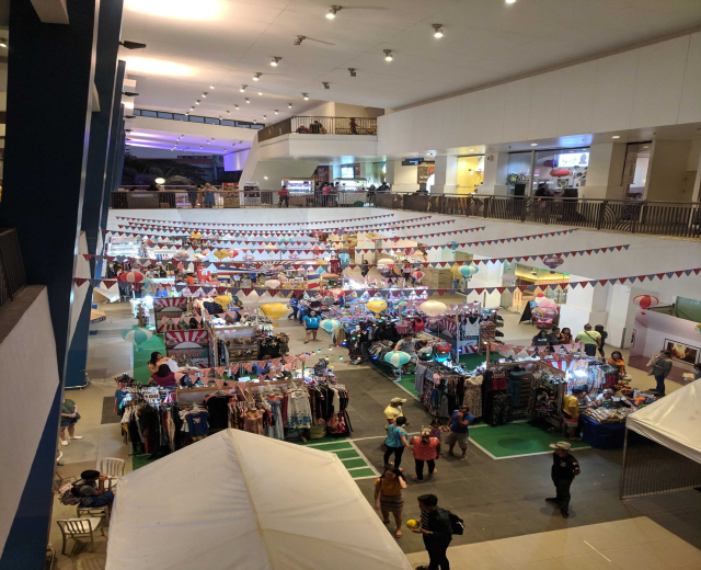 Ein volles Einkaufszentrum mit Kleiderständen, Dekorationsfahnen, Ballons und Menschen, die zwischen Geländern, Säulen und Deckenlampen sitzen und stehen, suggeriert einen Weihnachtsmarkt im Mall of Asia.