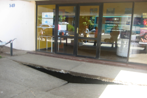 Eine gebrochene Oberfl√§che mit einer Glast√ľr, Wand und Stange im Hintergrund, die sich bewegende Fahrzeuge und einige stehende Personen reflektiert.