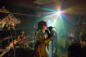 Eine Frau in einer schwarzen Jacke singt in ein Mikrofon auf der Bühne, mit einem Mann in Sonnenbrille und einem Stirnband, der ein Instrument spielt, und einem Plakat mit der Aufschrift "LA Guns" im Hintergrund.