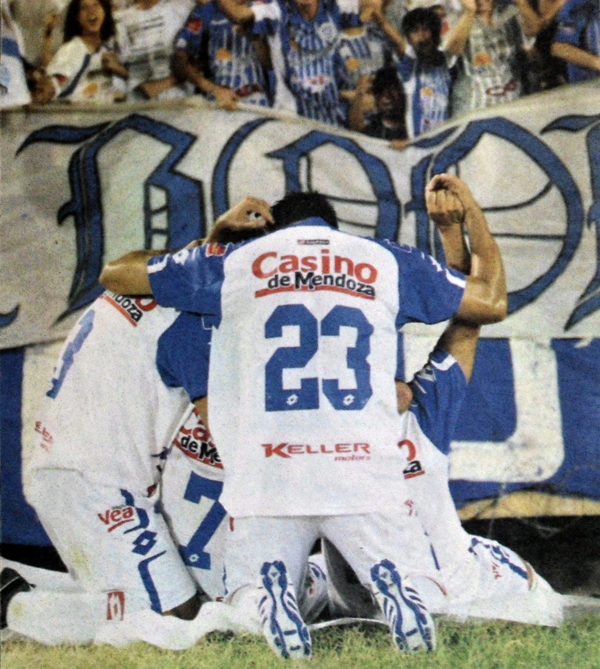 Gruppe von Fußballspielern, die auf dem Boden vor einer jubelnden Menge feiern, mit einem Banner im Hintergrund.